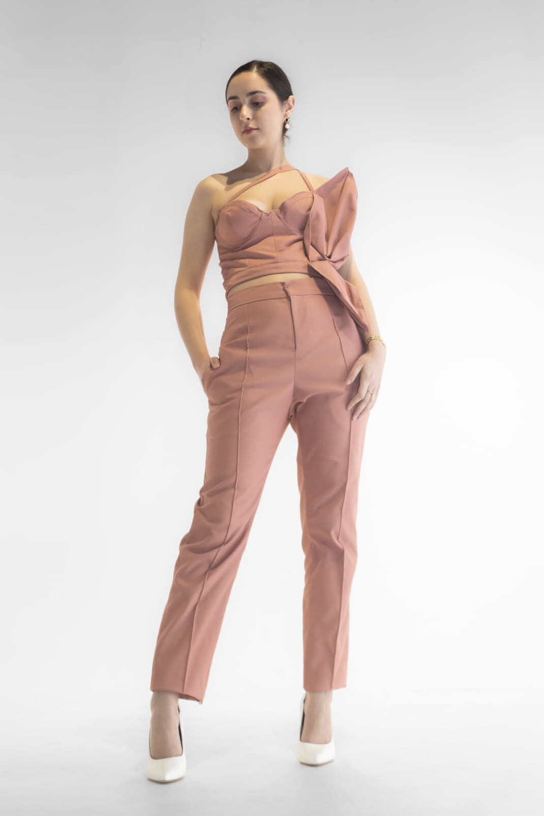 A woman models a sophisticated peach-colored jumpsuit with a structured bodice and asymmetrical peplum detail, paired with white heels.
