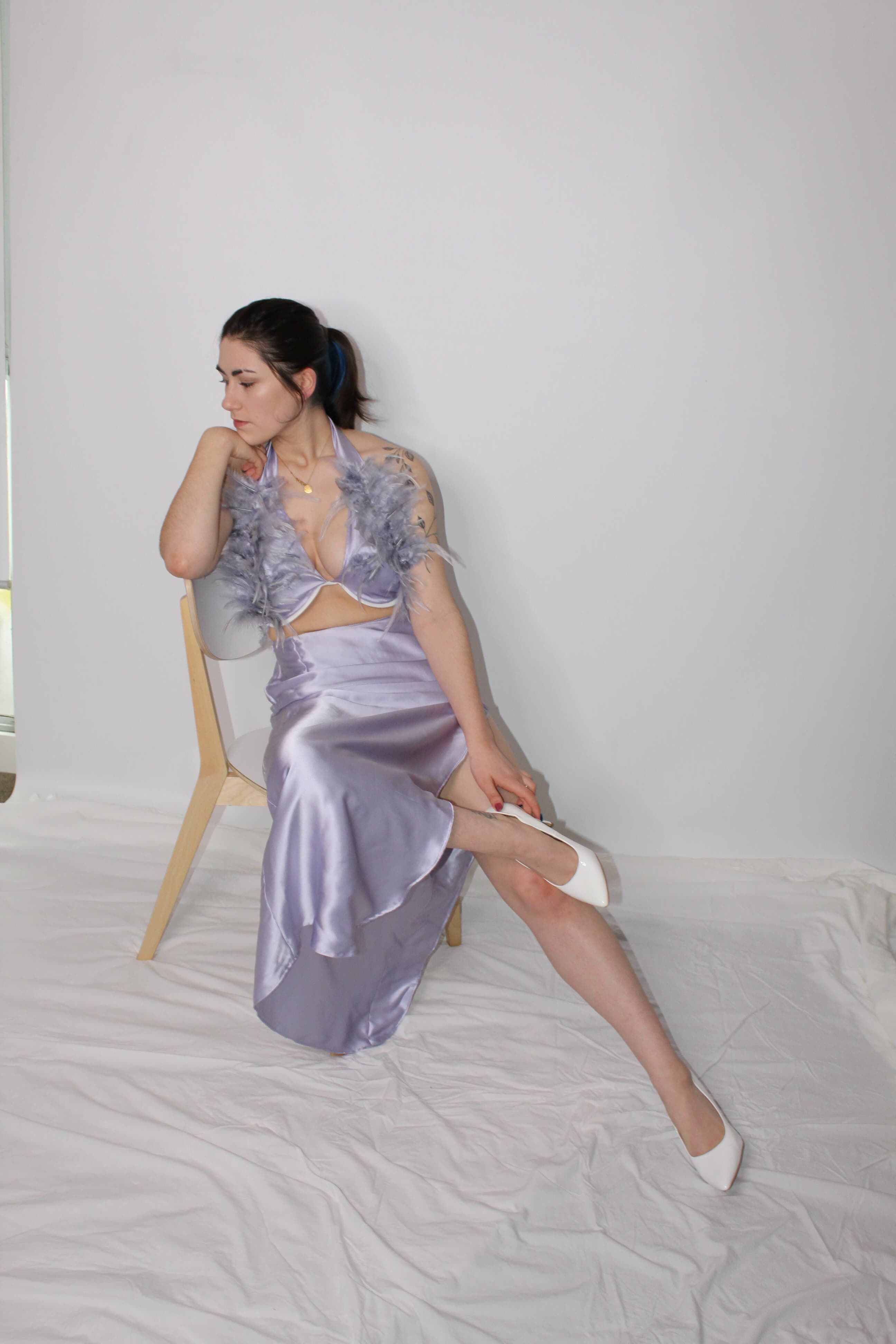 A woman seated on a wooden chair, donning a lavender dress with feather accents, adjusting her white heel against a plain backdrop.