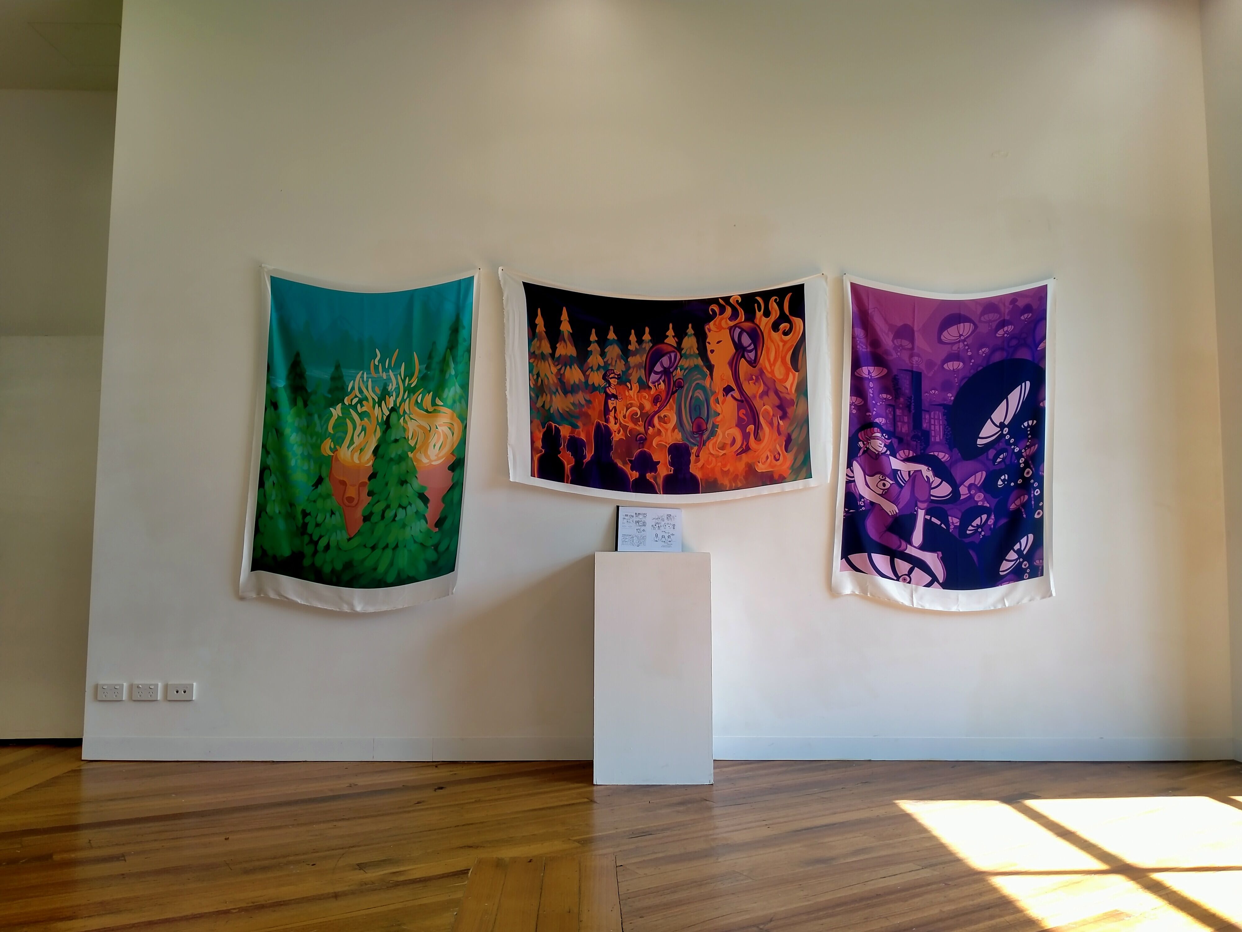 Three colorful tapestries depicting abstract and mythical themes displayed on a gallery wall, illuminated by natural light.
