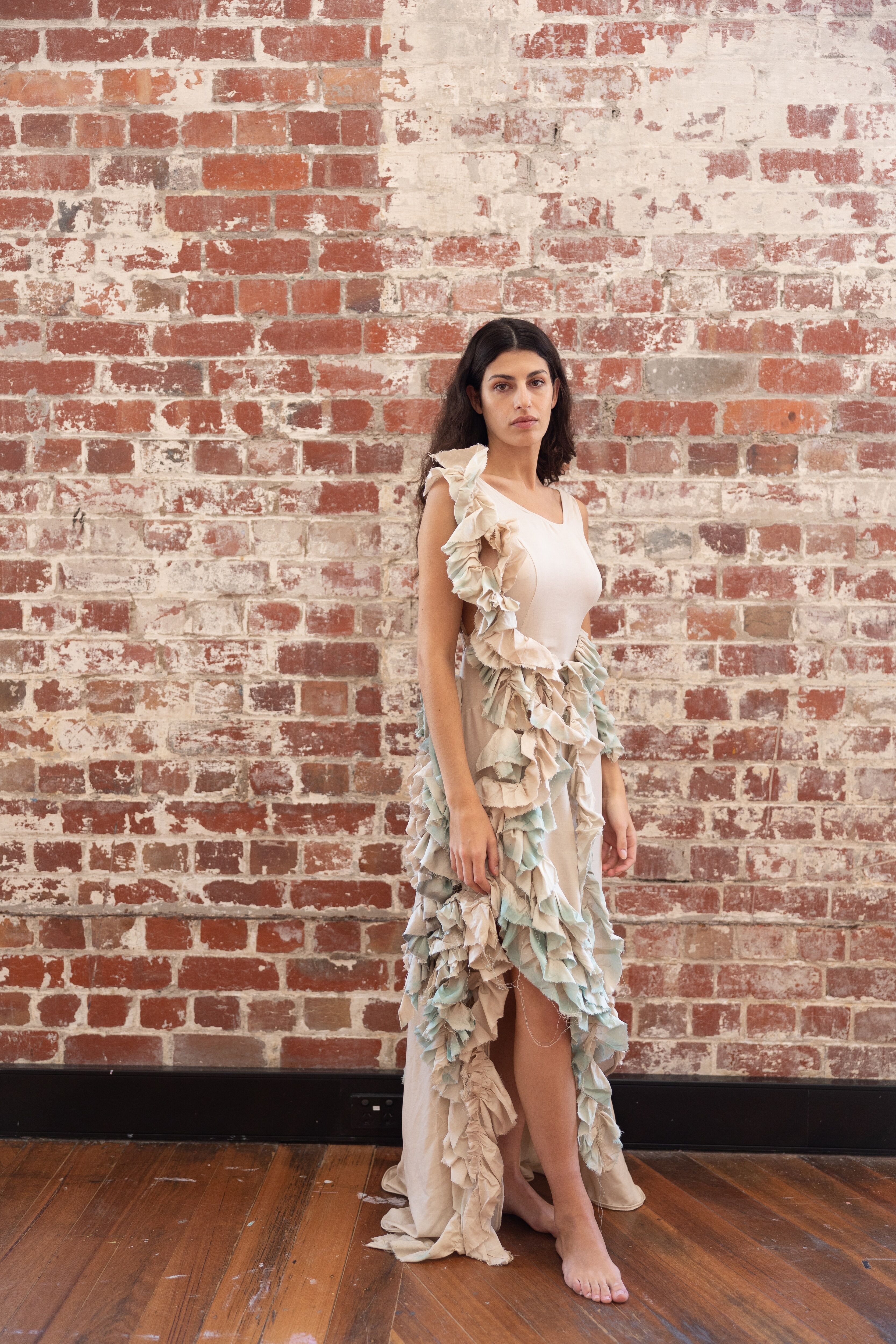 A poised woman stands against a rustic brick wall, her flowing dress adorned with delicate, petal-like ruffles, embodying a graceful juxtaposition of urbanity and natural forms.