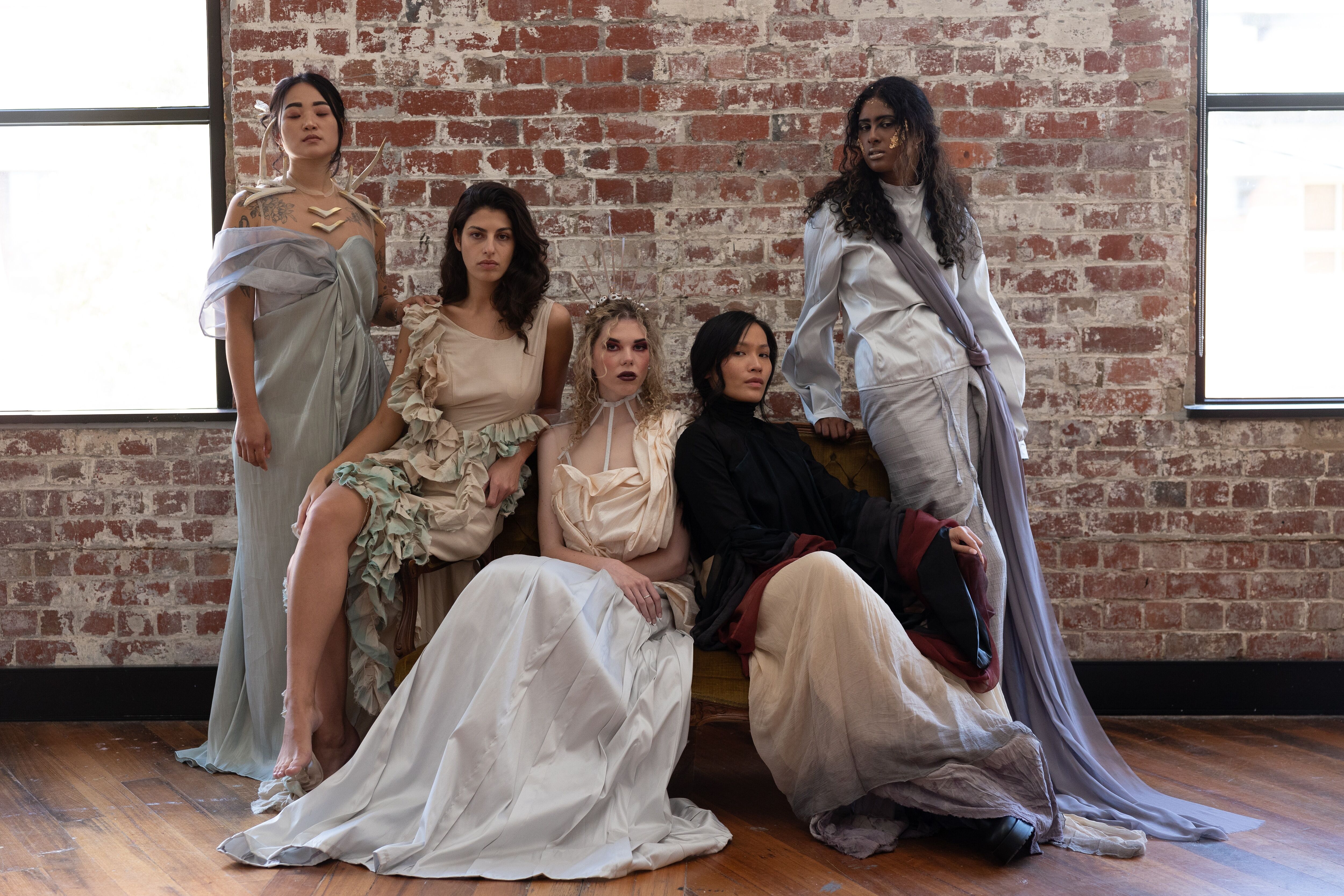 Five models posing in a rustic loft, each adorned in unique, flowy avant-garde attire, exuding an air of contemporary elegance against the exposed brick backdrop.