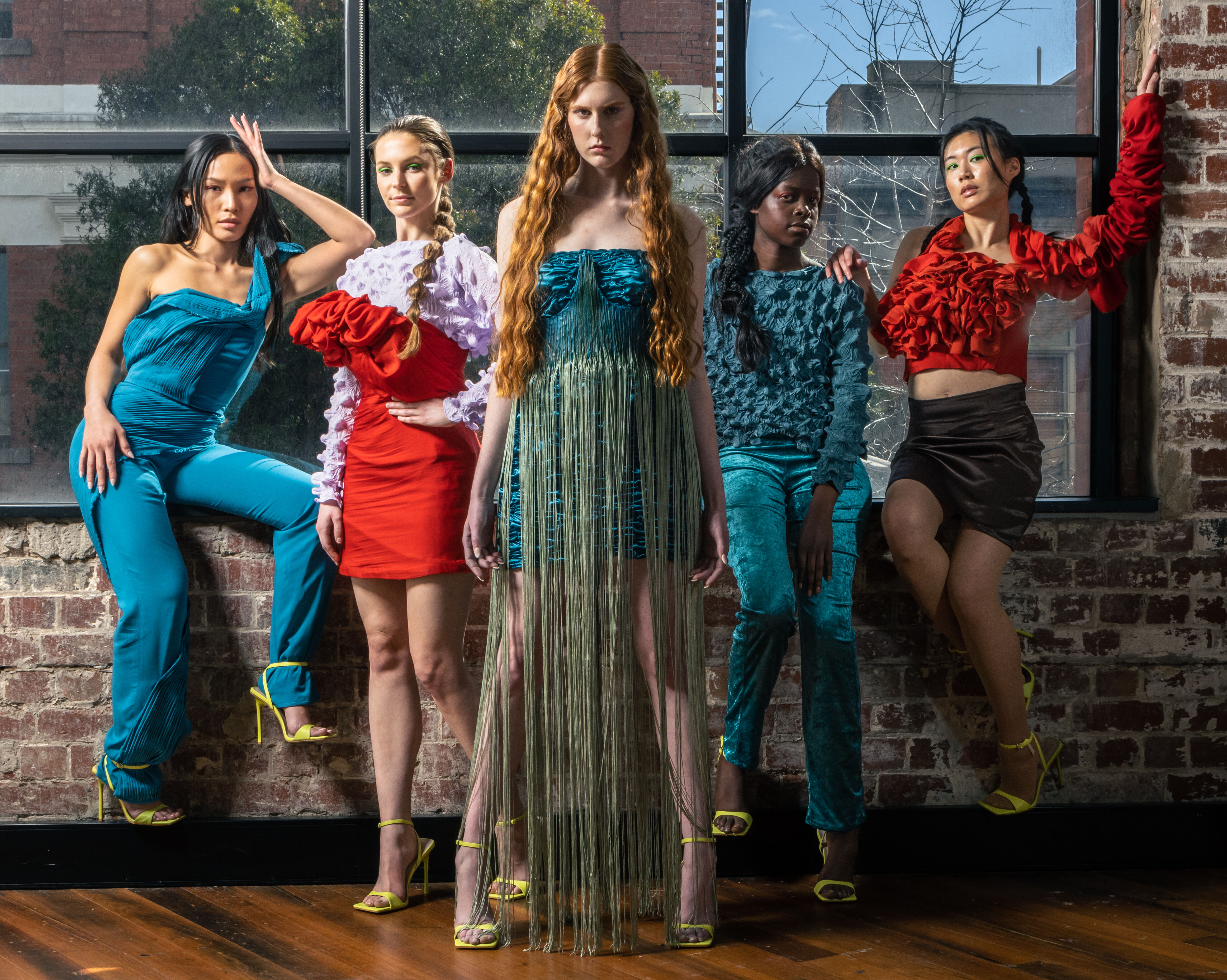 Five models in eclectic attire pose confidently in a loft, showcasing a blend of bold colors and textures against a rustic brick backdrop.