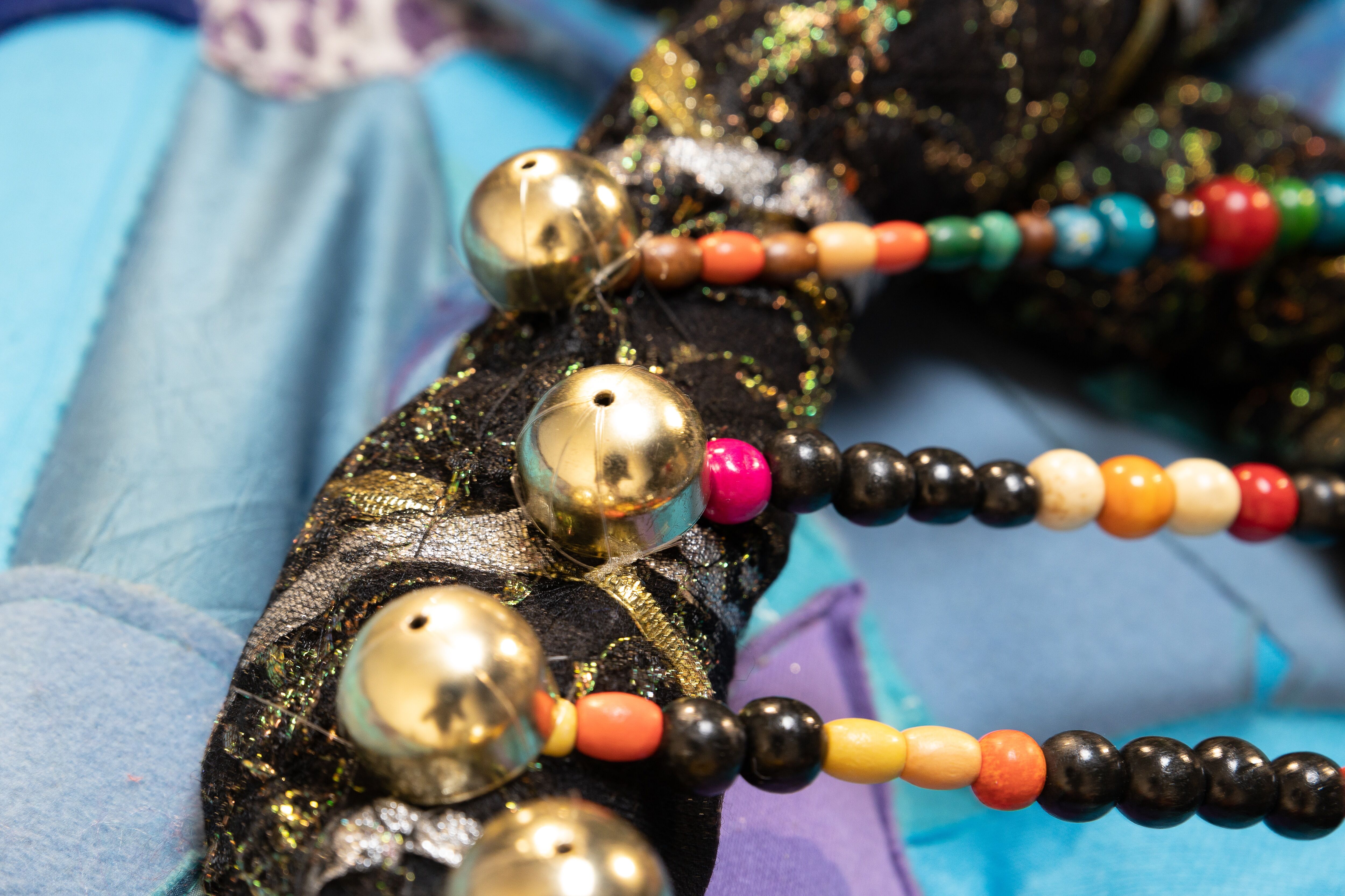 A close-up of multicolored beads and golden baubles arranged on glittery black fabric with hints of blue and purple textiles.