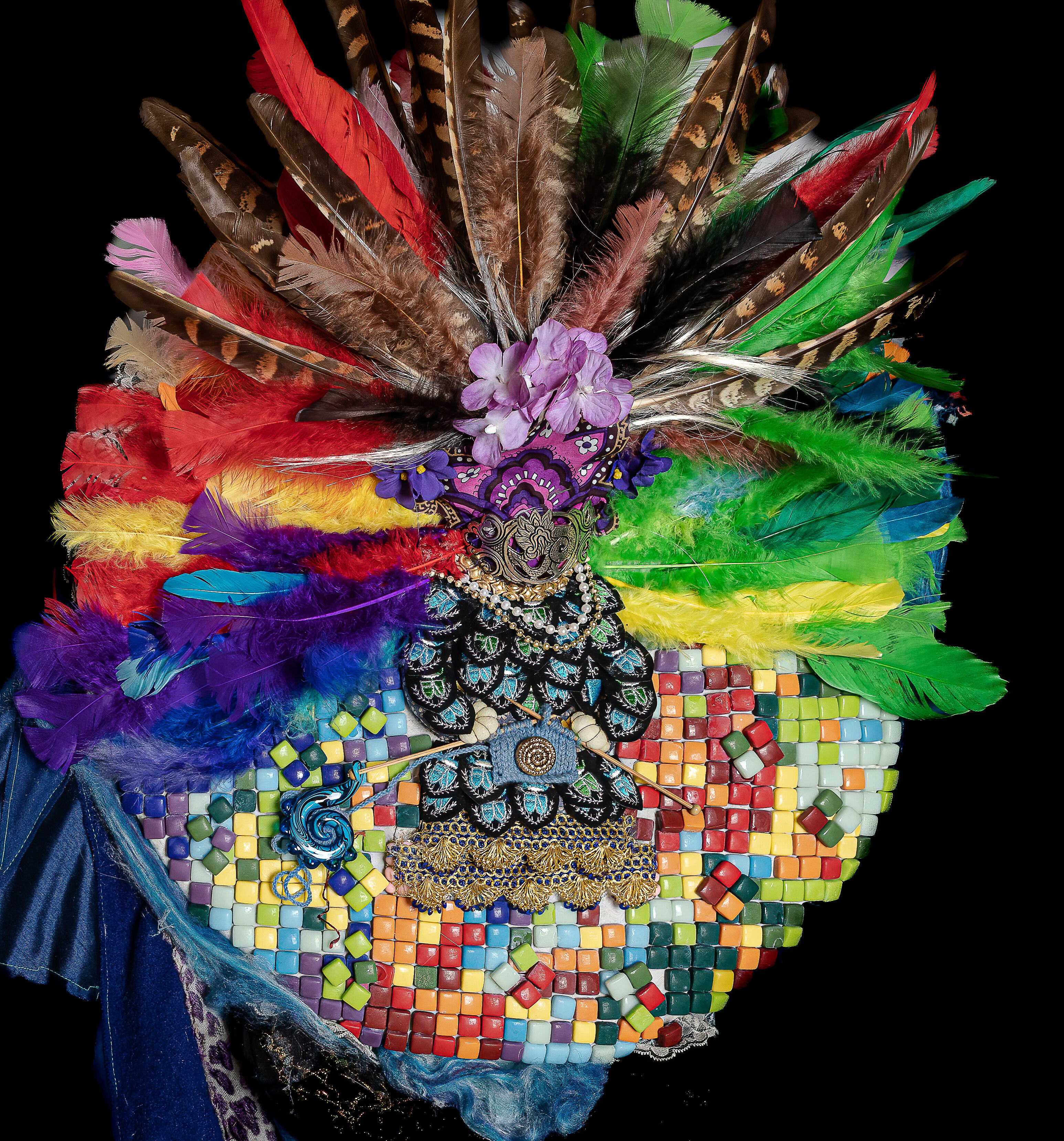 A vibrant carnival headdress featuring a spectrum of colorful feathers, intricate beadwork, and mosaic details on a black background.