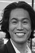 A cheerful young man with shoulder-length wavy hair, smiling broadly in a black and white photo.