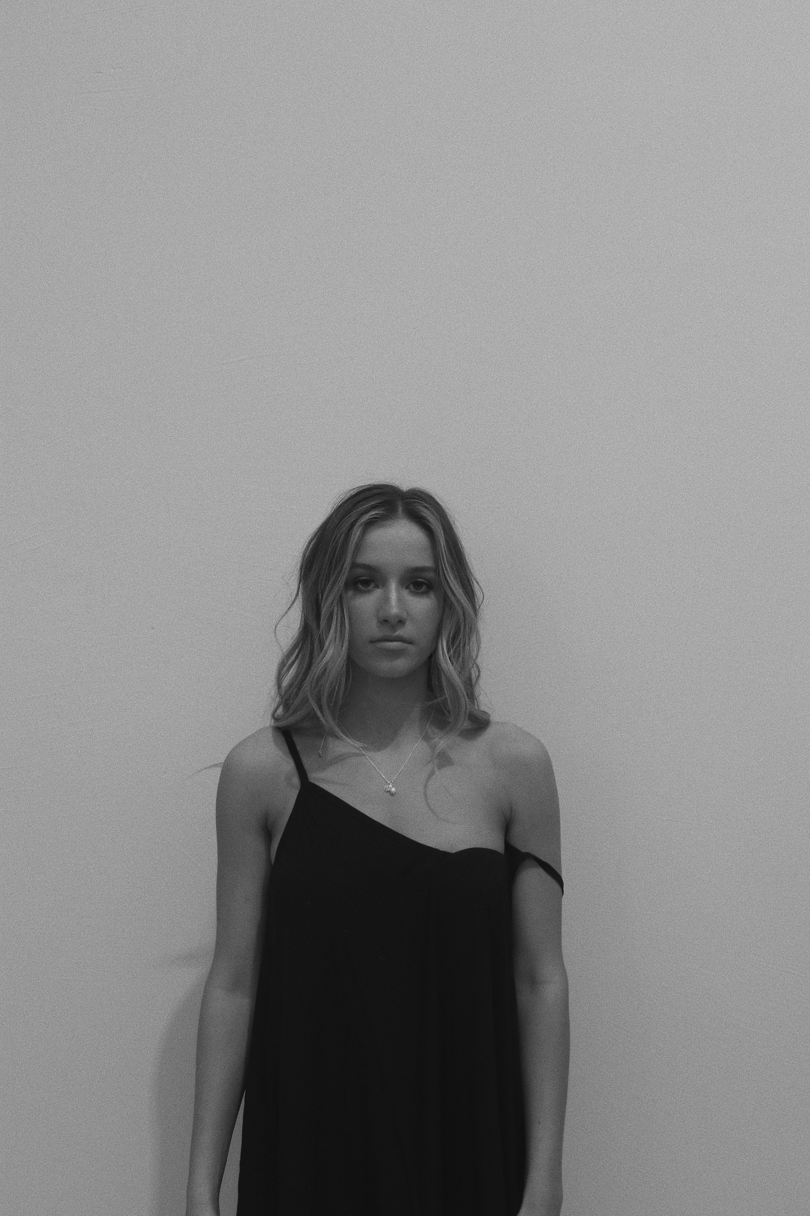 A grayscale image capturing a young woman in a black dress, standing against a plain background, her expression pensive.
