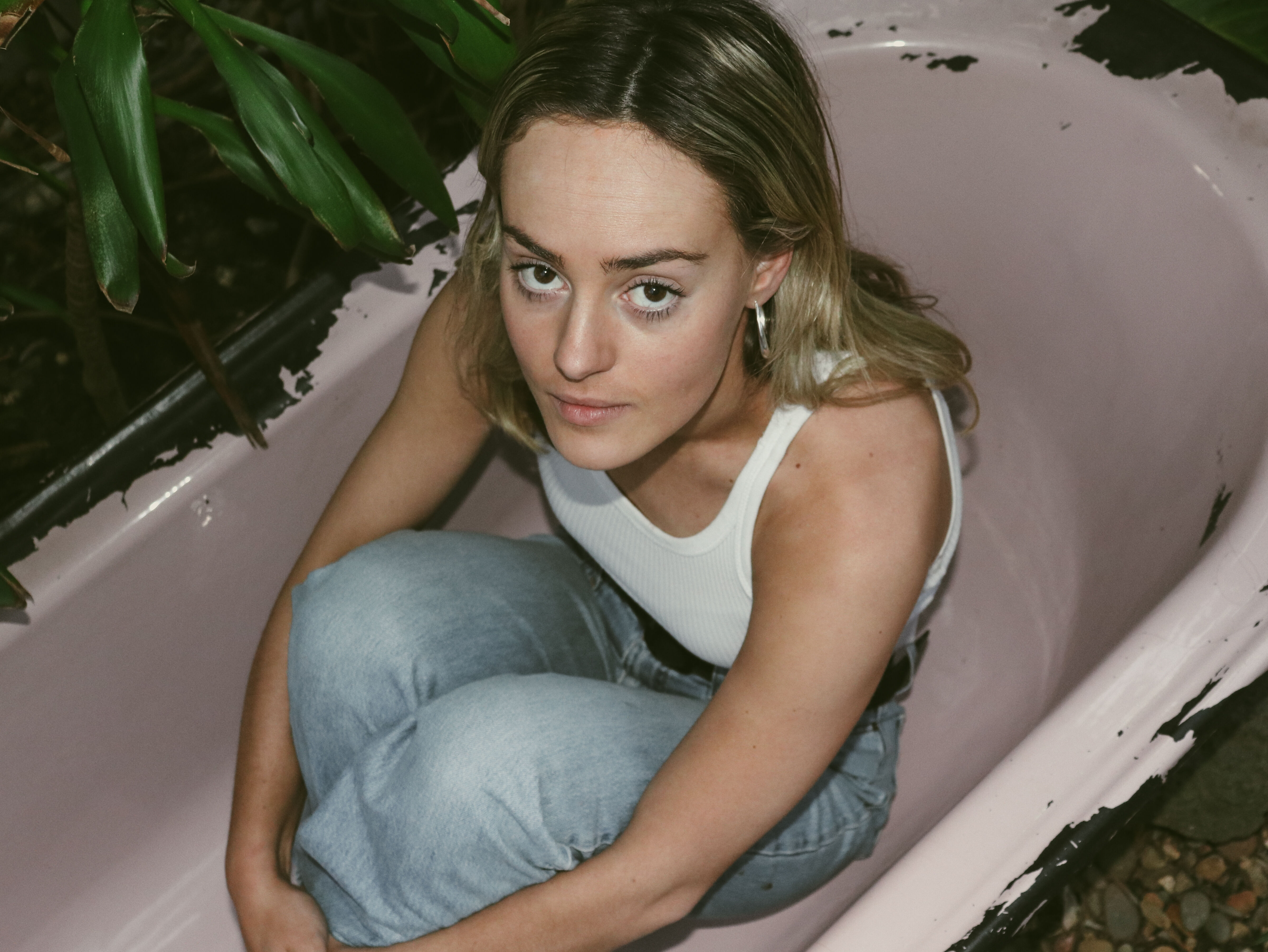 A young woman with blonde hair sits thoughtfully in an empty bathtub, surrounded by green foliage.