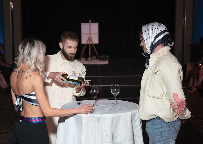 A scene captures a model serving wine to another, adding a theatrical twist to a fashion event.