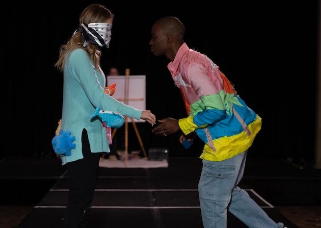 Two models engage in a playful interaction on stage, dressed in colorful, imaginative outfits.