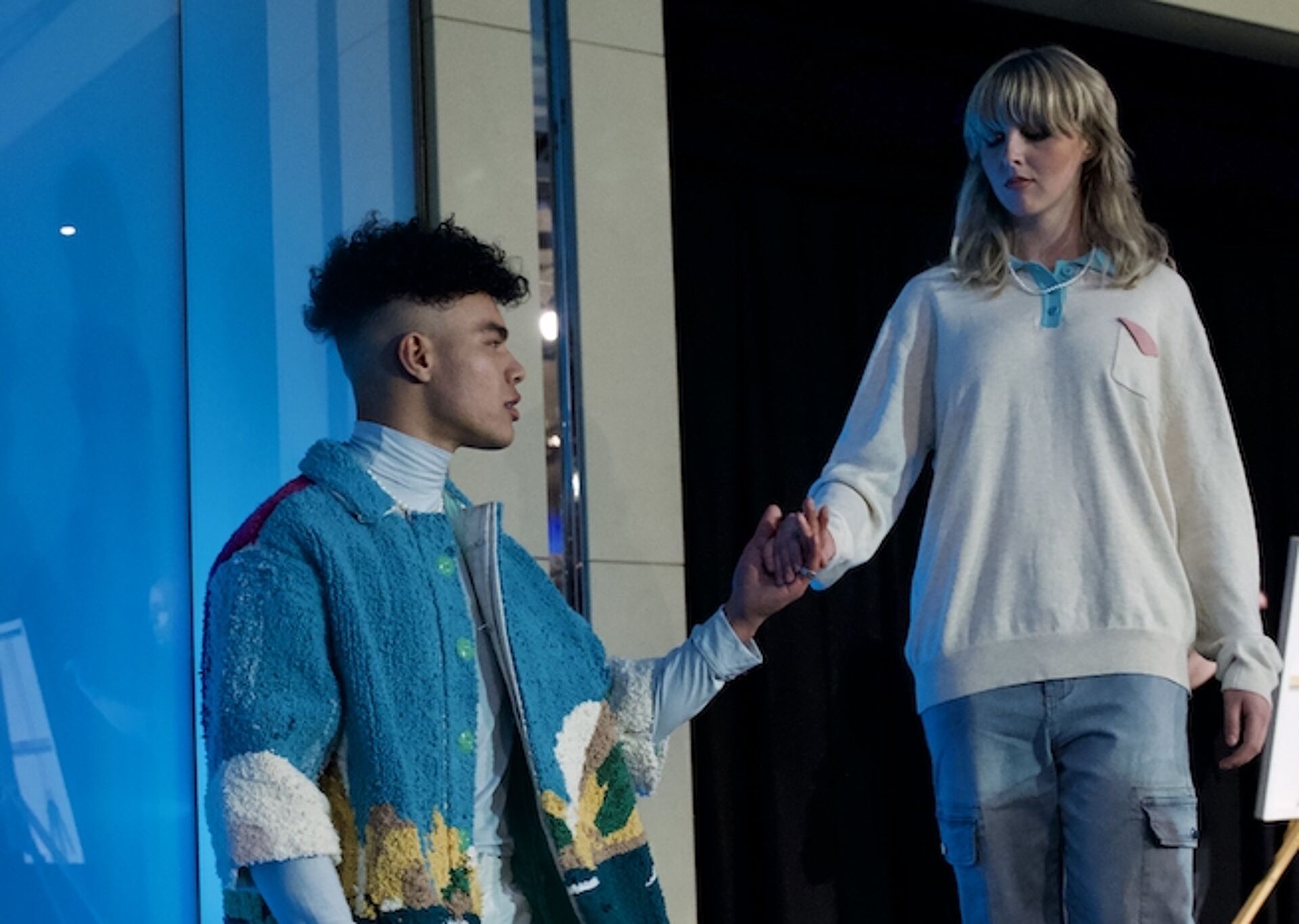 Two models connect in a dynamic pose, showcasing modern casual wear with a colorful, textured jacket and relaxed sweater.