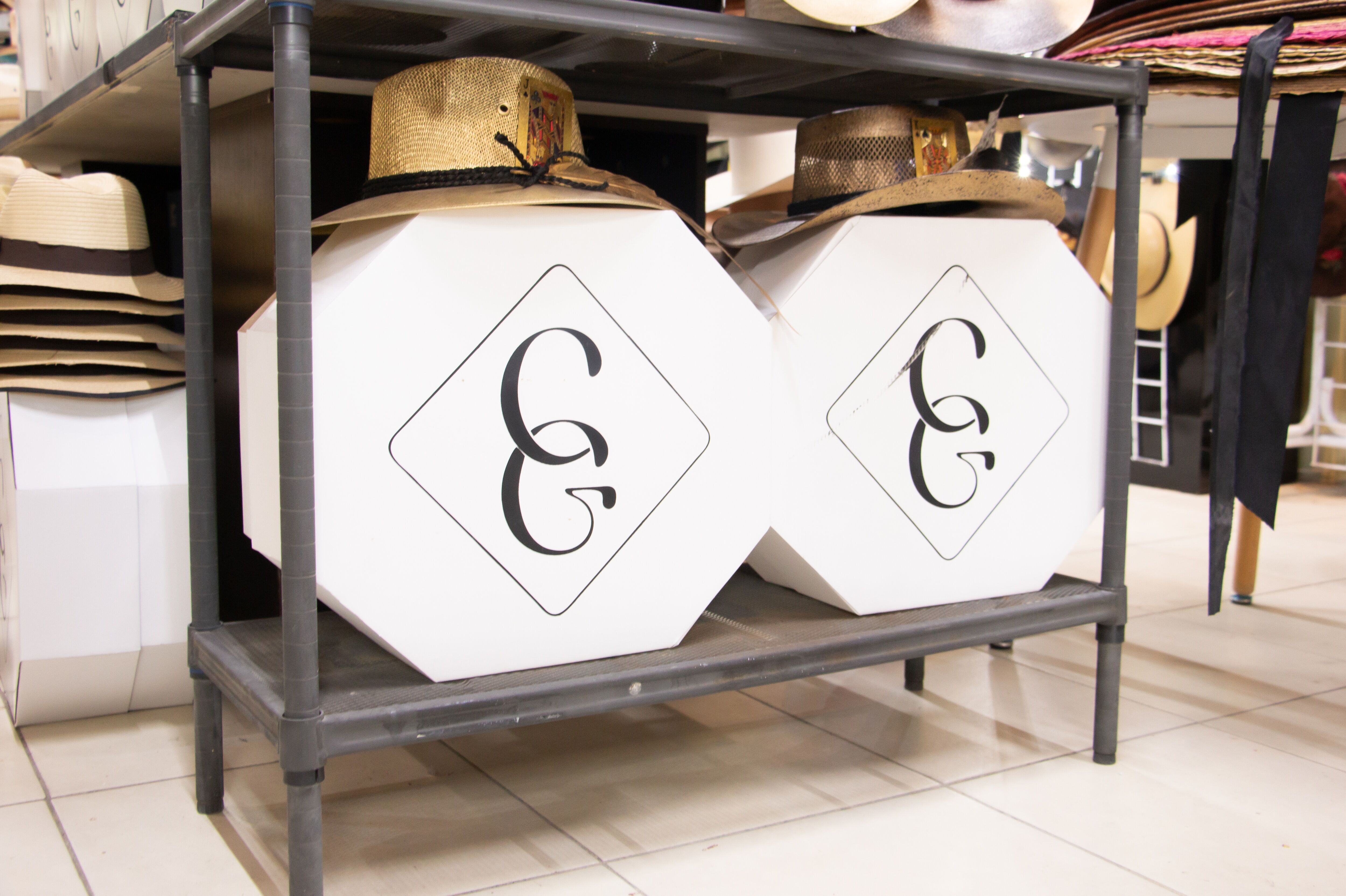 Una estantería ordenada en una tienda, mostrando varios estilos de sombreros dispuestos encima y cajas con logotipos de monograma estilosos debajo.