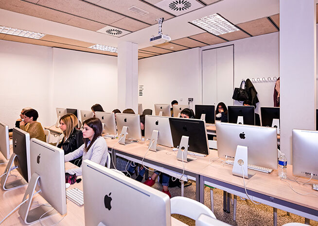 La imagen muestra un laboratorio de computadoras moderno con filas de estudiantes sentados en escritorios, cada uno trabajando en una computadora Apple iMac. El laboratorio tiene un diseño simple con paredes blancas, iluminación fluorescente y un proyector montado en el techo. En el fondo, un perchero sostiene bolsas y abrigos, indicando que los estudiantes están estudiando en un entorno académico.