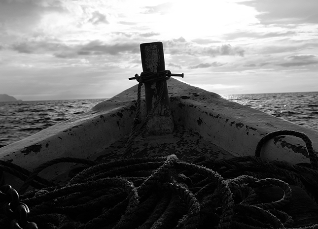  Imagen en blanco y negro de la proa de un barco con cuerda enrollada, frente a un mar tranquilo y cielo nublado.