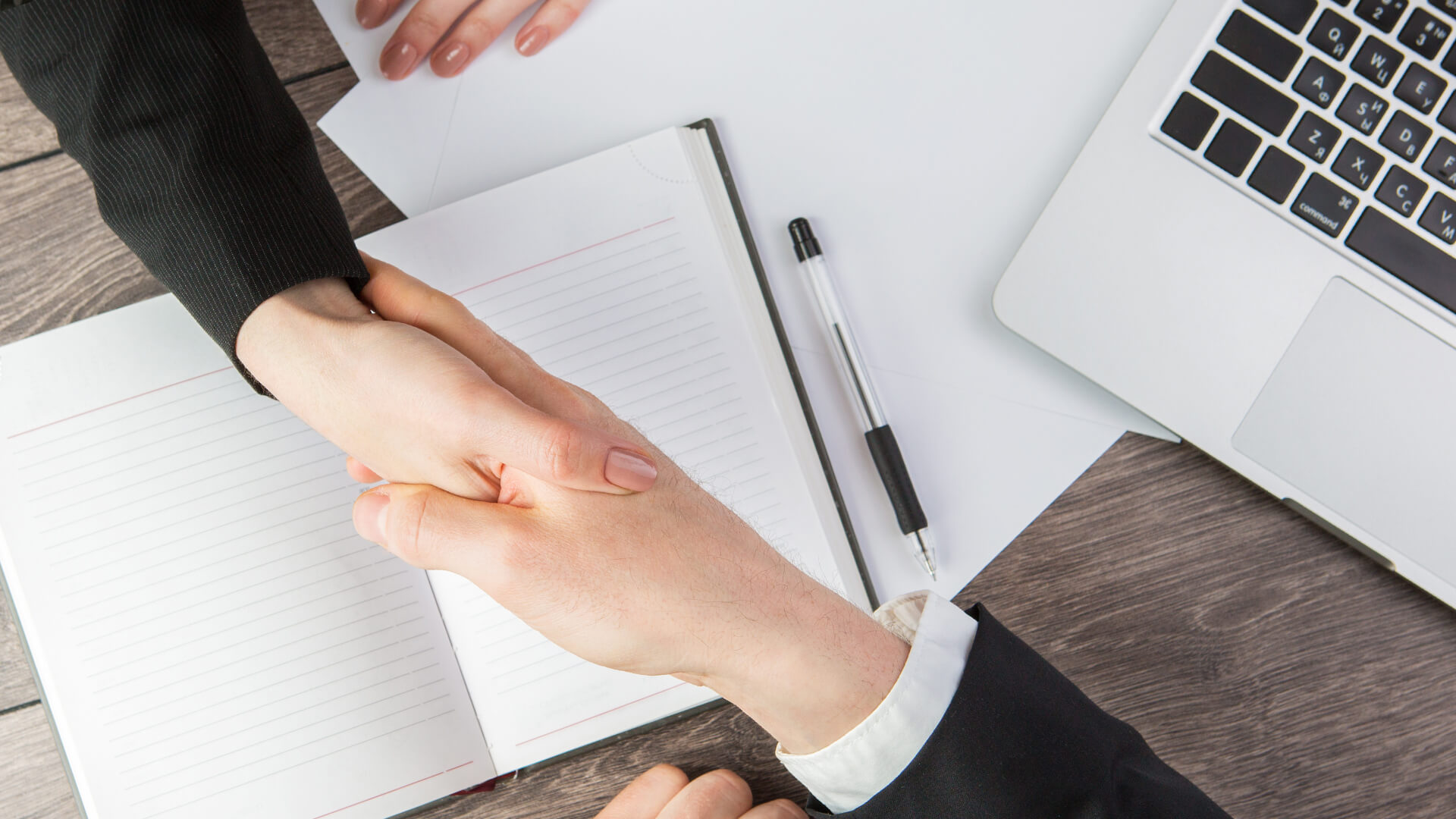 Two professionals engage in a firm handshake above a desk with an open notebook, pen, and a laptop, symbolizing agreement or partnership.