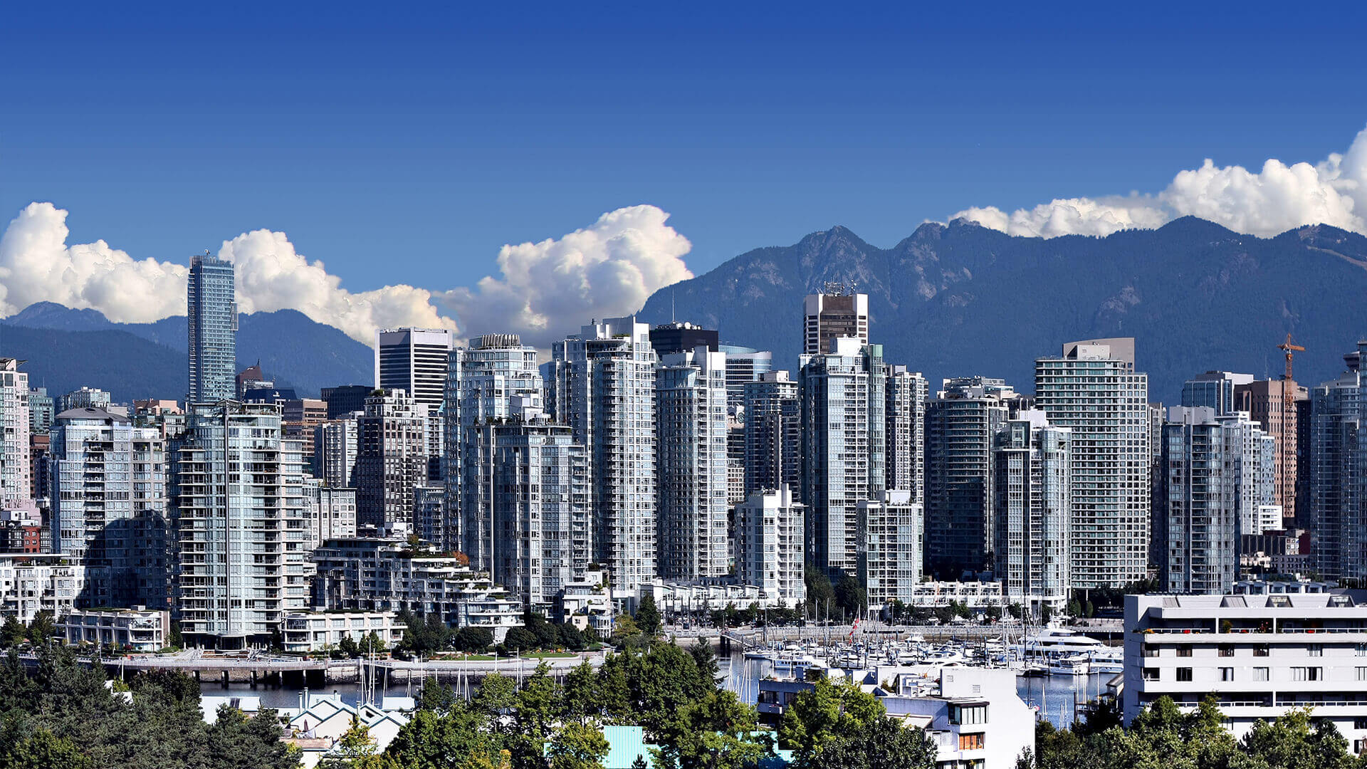 A panoramic view of Vancouver's dense urban skyline with residential and commercial high-rises against a backdrop of the majestic North Shore Mountains and scattered cumulus clouds.